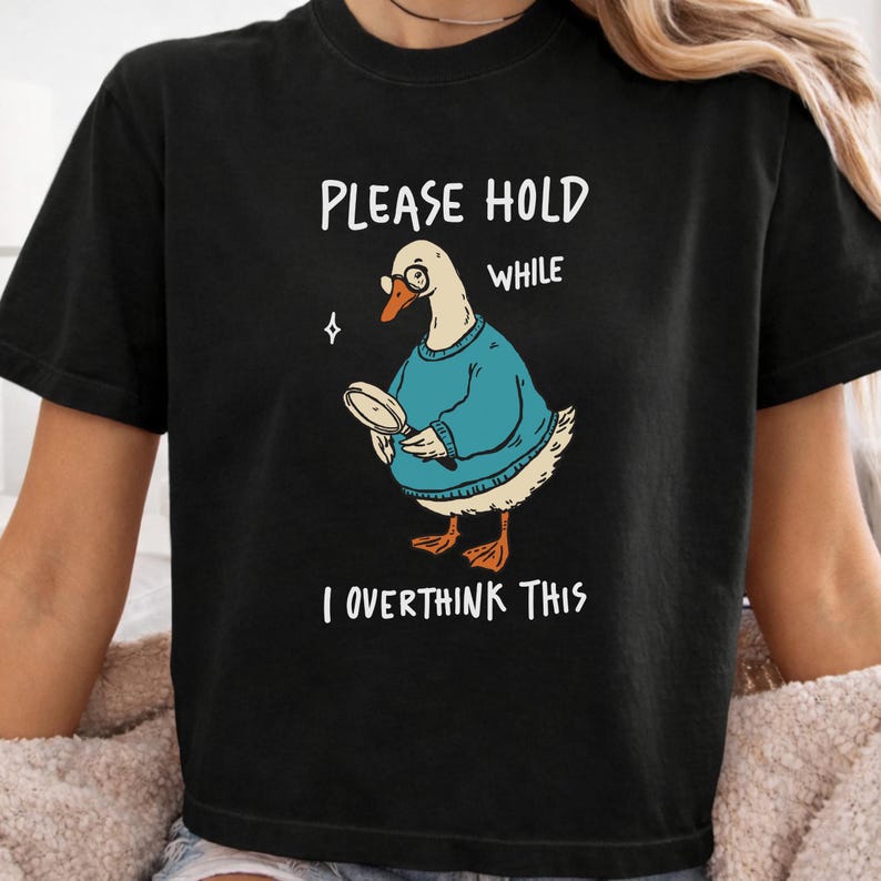 May include: A black t-shirt featuring a graphic of a white goose wearing a blue sweater and glasses, holding a magnifying glass. The text reads: PLEASE HOLD WHILE I OVERTHINK THIS. The design includes a small white star icon near the goose's head.