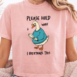 May include: A light pink t-shirt featuring a graphic of a white duck wearing a blue sweater and glasses while holding a magnifying glass. The text above the duck reads "PLEASE HOLD" and the text below the duck reads "WHILE I OVERTHINK THIS".