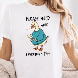 May include: A white short-sleeved t-shirt featuring a graphic of a goose wearing glasses and a teal sweater, holding a magnifying glass. The text above and below the goose reads, "PLEASE HOLD WHILE I OVERTHINK THIS" in black, hand-drawn style lettering.