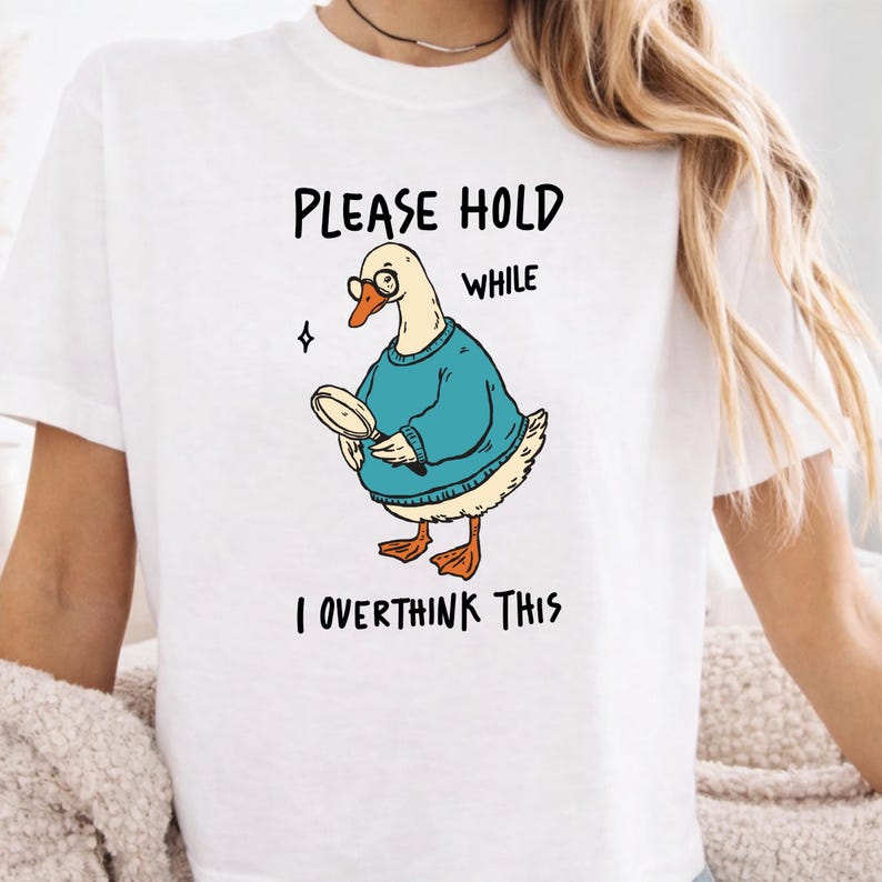 May include: A white short-sleeved t-shirt featuring a graphic of a goose wearing glasses and a teal sweater, holding a magnifying glass. The text above and below the goose reads, "PLEASE HOLD WHILE I OVERTHINK THIS" in black, hand-drawn style lettering.