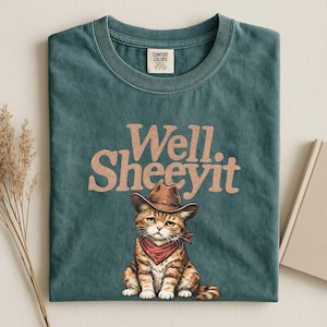 May include: Teal Comfort Colors t-shirt featuring a cartoon cat wearing a cowboy hat and red bandana. The shirt has the text "Well. Sheeeyit" in a brown, retro font. The shirt is folded neatly.