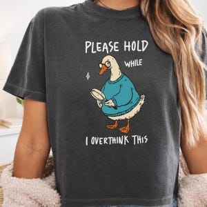 May include: A charcoal gray t-shirt featuring a graphic of a white goose wearing round glasses and a blue sweater, holding a magnifying glass. The text reads: PLEASE HOLD WHILE I OVERTHINK THIS. The design includes a small four-pointed star icon.