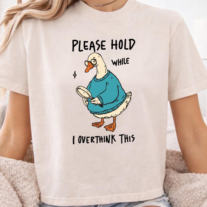 May include: A cream-colored t-shirt featuring a graphic of a white goose wearing a blue sweater and round glasses, holding a magnifying glass. The text reads, PLEASE HOLD WHILE I OVERTHINK THIS, in black lettering above and below the illustration.