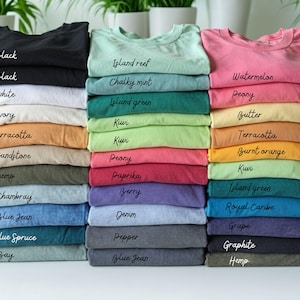 May include: A collection of folded t-shirts stacked in three columns, featuring a variety of colors including black, white, ivory, terracotta, sandstone, hemp, chambray, blue jean, blue spruce, bay, island reef, chalky mint, island green, kiwi, peony, paprika, berry, denim, pepper, watermelon, butter, burnt orange, royal caribe, grape, and graphite, with each color name printed on the fabric.
