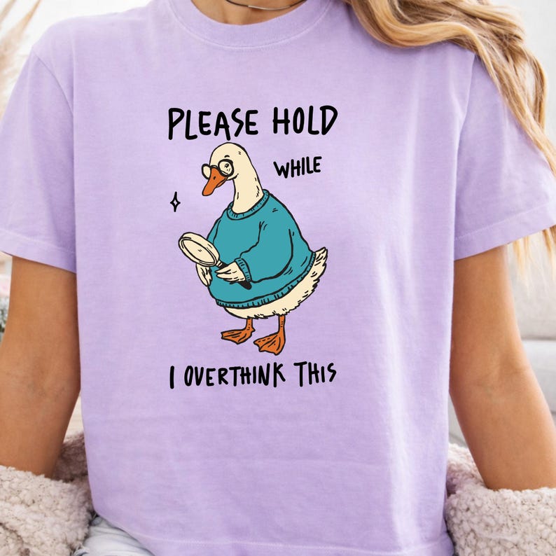 May include: A light purple t-shirt featuring a graphic of a white goose wearing round glasses and a teal sweater, holding a magnifying glass. The text reads: PLEASE HOLD WHILE I OVERTHINK THIS. The design includes a small four-pointed star icon.