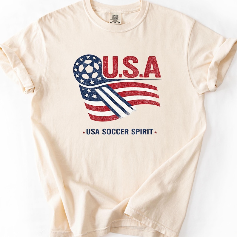Puede incluir: Una camiseta de manga corta color crema con un gr&aacute;fico de un bal&oacute;n de f&uacute;tbol integrado en una bandera estadounidense ondeante en rojo, blanco y azul. El texto U.S.A. aparece sobre el gr&aacute;fico y el texto USA SOCCER SPIRIT est&aacute; impreso debajo.