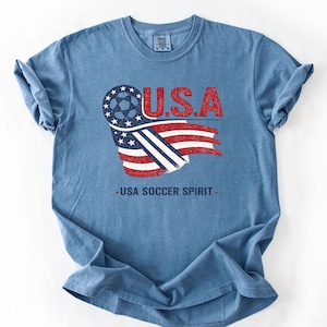 Puede incluir: Una camiseta azul de manga corta con un gr&aacute;fico de un bal&oacute;n de f&uacute;tbol integrado en un dise&ntilde;o de bandera estadounidense. El texto U.S.A. aparece en rojo sobre el gr&aacute;fico, y el texto USA SOCCER SPIRIT est&aacute; impreso en azul oscuro debajo.
