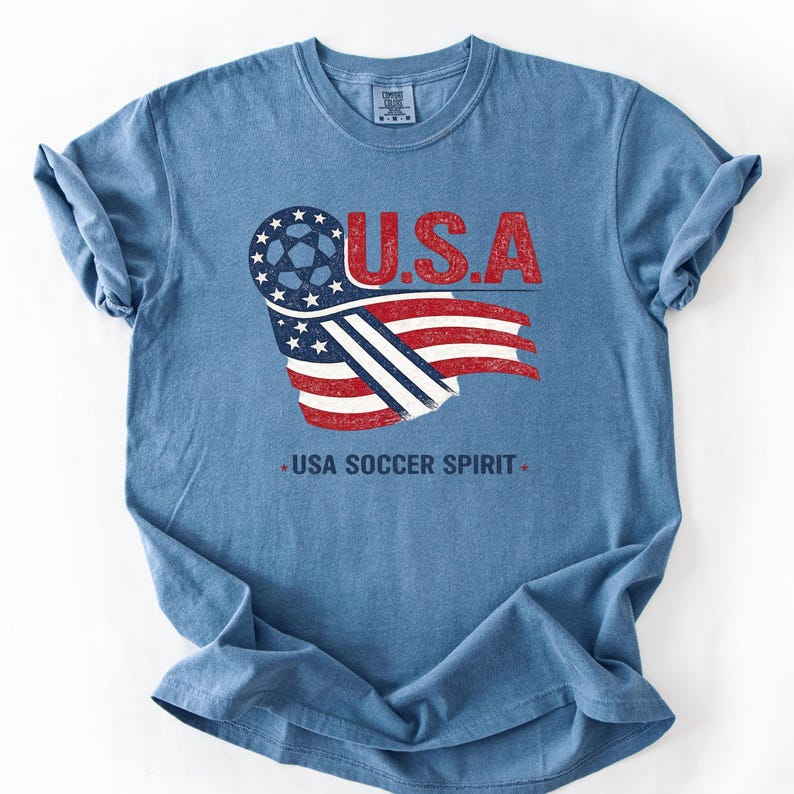 Puede incluir: Una camiseta azul de manga corta con un gr&aacute;fico de un bal&oacute;n de f&uacute;tbol integrado en un dise&ntilde;o de bandera estadounidense. El texto U.S.A. aparece en rojo sobre el gr&aacute;fico, y el texto USA SOCCER SPIRIT est&aacute; impreso en azul oscuro debajo.