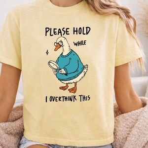May include: A pale yellow t-shirt featuring a graphic of a white goose wearing round glasses and a blue sweater. The goose holds a magnifying glass. The text reads: PLEASE HOLD WHILE I OVERTHINK THIS. The design is centered on the front of the shirt.