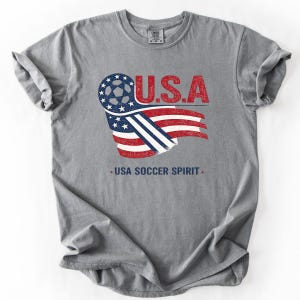Puede incluir: Una camiseta gris de manga corta con un dise&ntilde;o gr&aacute;fico que incluye el texto U.S.A. y USA SOCCER SPIRIT. El gr&aacute;fico muestra un bal&oacute;n de f&uacute;tbol integrado en una bandera estadounidense estilizada en colores rojo, blanco y azul con textura desgastada.