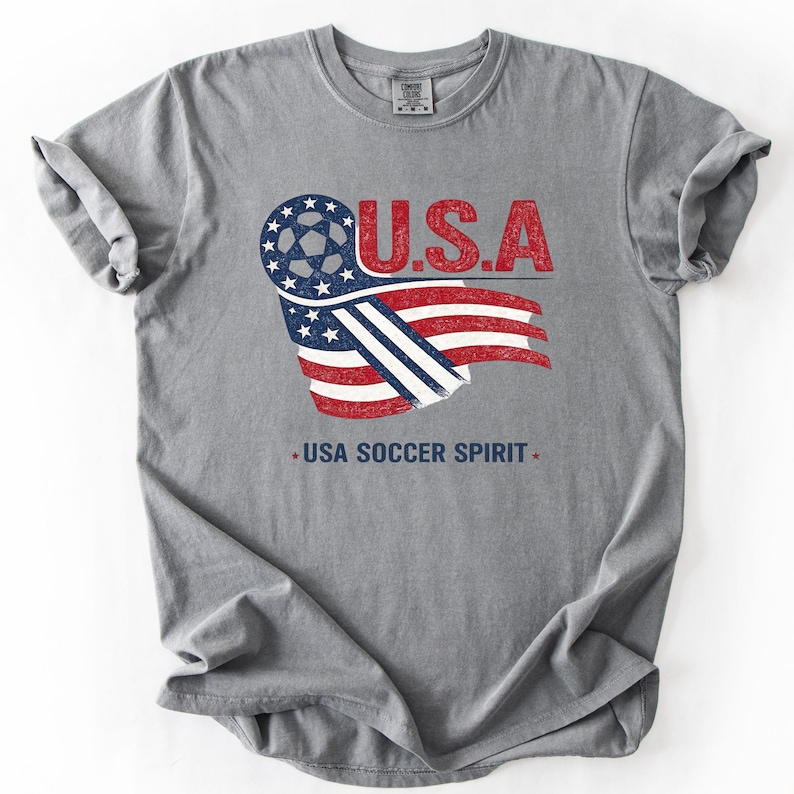 Puede incluir: Una camiseta gris de manga corta con un dise&ntilde;o gr&aacute;fico que incluye el texto U.S.A. y USA SOCCER SPIRIT. El gr&aacute;fico muestra un bal&oacute;n de f&uacute;tbol integrado en una bandera estadounidense estilizada en colores rojo, blanco y azul con textura desgastada.