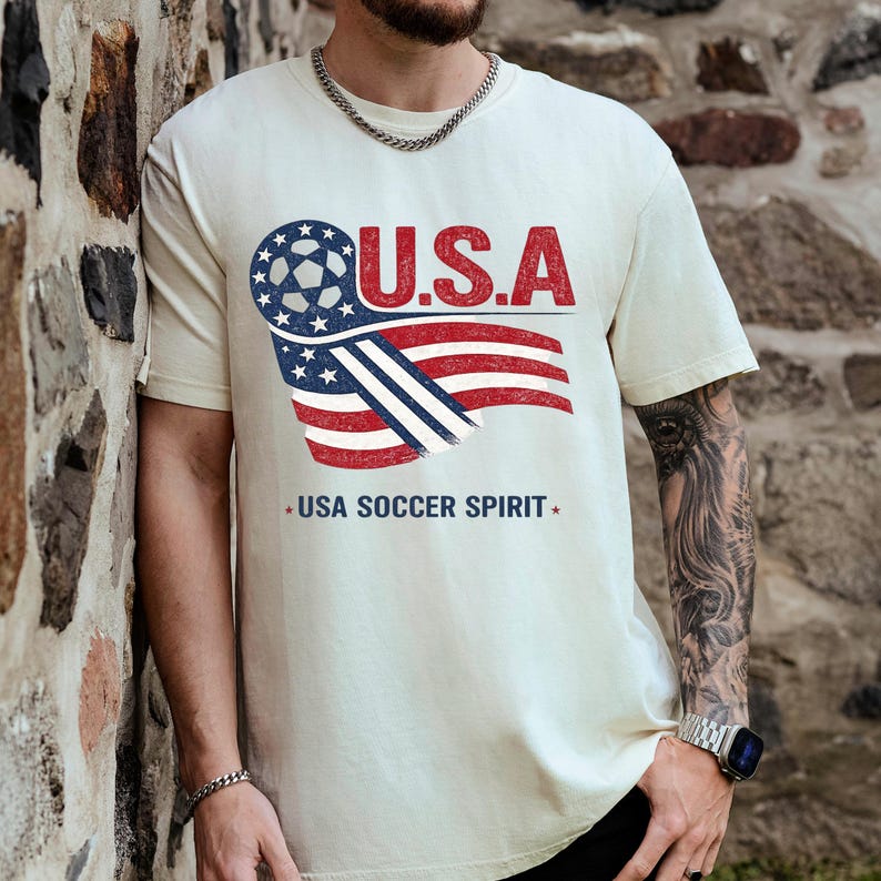 Puede incluir: Una camiseta de manga corta color crema con un dise&ntilde;o gr&aacute;fico que incluye el texto U.S.A. sobre un bal&oacute;n de f&uacute;tbol estilizado y un motivo de bandera estadounidense ondeante. Debajo del gr&aacute;fico, el texto USA SOCCER SPIRIT est&aacute; impreso en azul.