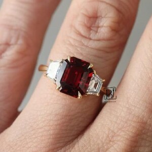 Puede incluir: Un anillo de oro con una gran piedra preciosa rectangular de color rojo intenso flanqueada por dos piedras blancas más pequeñas en forma de trapecio. El anillo está en un dedo, mostrando el diseño y el contraste de color.
