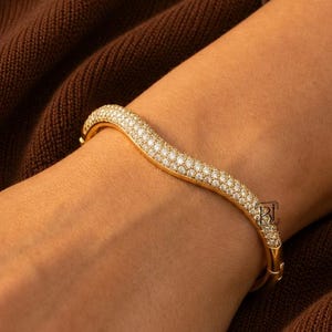 May include: A gold-toned hinged bangle bracelet featuring a wavy, serpentine silhouette. The surface of the metal is densely encrusted with numerous small, clear, sparkling stones that cover the entire visible exterior of the curved band.