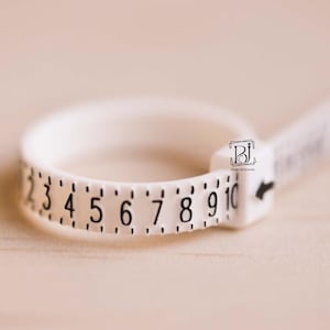May include: A white ring sizer tool with black numbers from 2 to 10. The tool is used to measure ring sizes. The tool is made of plastic and has a sliding mechanism to adjust the size. The background is a light brown color.