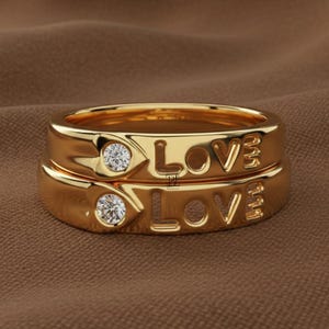 May include: Two stacked gold rings, each featuring a polished band with a circular, clear, faceted gemstone set into a heart-shaped frame on the left side, followed by the word LOVE embossed in capital letters on the right side of the band.