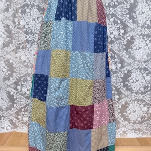 Puede incluir: Una falda larga hasta el suelo confeccionada con un mosaico de cuadrados de tela. El tejido presenta una gran variedad de estampados florales y geométricos en tonos azul, verde, oliva, burdeos y beige, con una cinturilla ajustada.