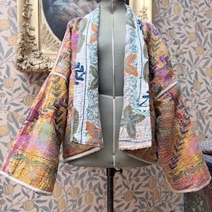 May include: A quilted jacket featuring a vibrant kantha-style embroidery with multi-coloured floral and vine patterns on a yellow and pink base. The garment has wide sleeves, an open front, and a white trim along the edges and lapels.