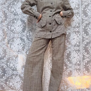 May include: A two-piece suit featuring a long-sleeved button-down jacket with a pointed collar, shoulder epaulets, and two front flap pockets, paired with matching wide-leg trousers, all in a brown and tan plaid check pattern.
