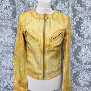 Könnte beinhalten: Senfgelbe Lederjacke mit Reißverschluss, zwei Reißverschlusstaschen auf der Brust und dekorativen Nähten. Die Jacke wird auf einer Schaufensterpuppe vor einem floral gemusterten Hintergrund präsentiert.