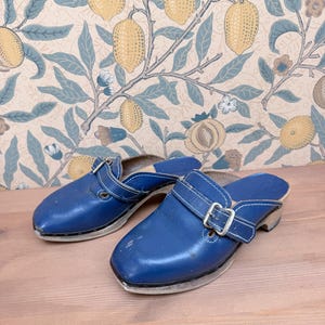 Pode incluir: Um par de tamancos de couro azul com fivelas prateadas e costuras brancas. Os tamancos têm sola de madeira e uma tira na parte superior. Fundo com papel de parede com tema de limões.