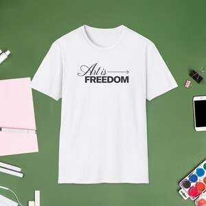 May include: A white short-sleeved t-shirt featuring the black text Art is FREEDOM with an arrow pointing to the right. The shirt has a crew neckline and a standard fit, displaying the graphic clearly centered on the chest area of the garment.