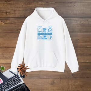 Puede incluir: Sudadera con capucha blanca con bolsillo delantero y un gráfico azul. El gráfico incluye las palabras "Artist Zone" e iconos de materiales de arte, una planta, auriculares, libros, una computadora y una taza de café. También se incluye el texto "Art Lives in the Little Things".