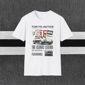 Może przedstawiać: Biała koszulka z grafiką Toyoty Supra, napisem "TOKYO AUTOS" i "THE ICONIC LEGEND". Projekt zawiera zdjęcia samochodu i słowo "PERFORMANCE".