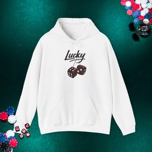 May include: White hoodie with a front pocket and drawstring. The word "Lucky" is written above a pair of dice. The dice are dark brown with white dots. The hoodie is a casual garment.