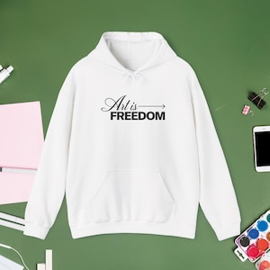 May include: A white long-sleeve hooded sweatshirt featuring a front kangaroo pocket and the black text "Art is FREEDOM" with an arrow graphic printed on the center chest. The garment has a drawstring hood and ribbed cuffs and hem.