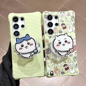 以下が含まれることがあります： 2つの薄緑色の携帯電話ケース。漫画のキャラクターデザイン。片方は青い帽子をかぶったキャラクター、もう片方は白いクマのようなキャラクター。どちらもポップアップグリップとカメラアレイを備えています。