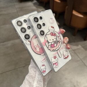 以下が含まれることがあります： ハローキティのデザインが施された透明な携帯電話ケース。ケースには、リボンとカップが付いたキャラクターなど、さまざまなピンクと白のハローキティのグラフィックが施されています。ケースはスマートフォンに合うように設計されています。