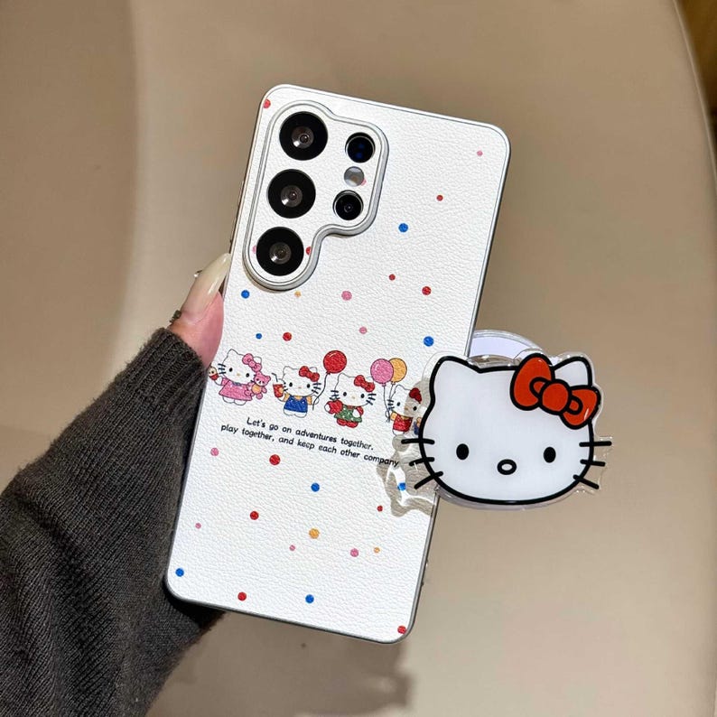 K&ouml;nnte beinhalten: Wei&szlig;es Handycase mit Hello Kitty-Design. Das Case hat ein sich wiederholendes Muster aus bunten Punkten und eine Reihe von Hello Kitty-Charakteren. Ein klarer Handygriff mit Hello Kitty-Gesicht und roter Schleife ist angebracht.
