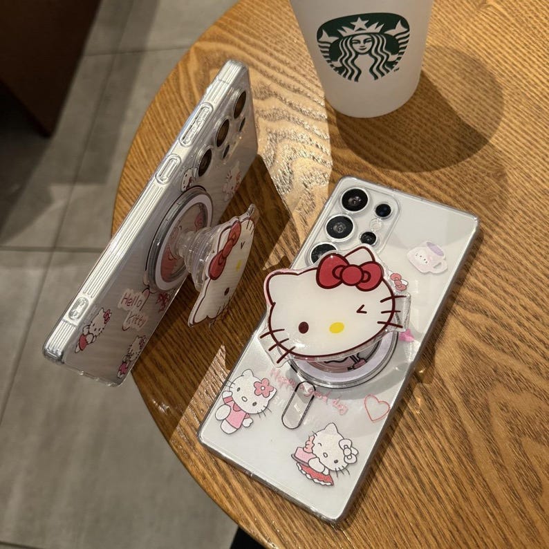 以下が含まれることがあります： ハローキティのデザインが施された透明な携帯電話ケース。1つのケースには、赤いリボンが付いたハローキティのポップアップグリップが付いています。ケースにはハローキティのグラフィックと「Hello Kitty」の文字が入っています。ケースは木製のテーブルの上にあります。