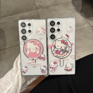 以下が含まれることがあります： ハローキティのデザインが施された透明な携帯電話ケース2つ。1つのケースには、ピンクのハローキティがピンクの円の中に描かれ、「Hello Kitty」の文字が入っています。もう1つのケースには、リボンと「Happy good day」の文字が入ったハローキティが描かれています。