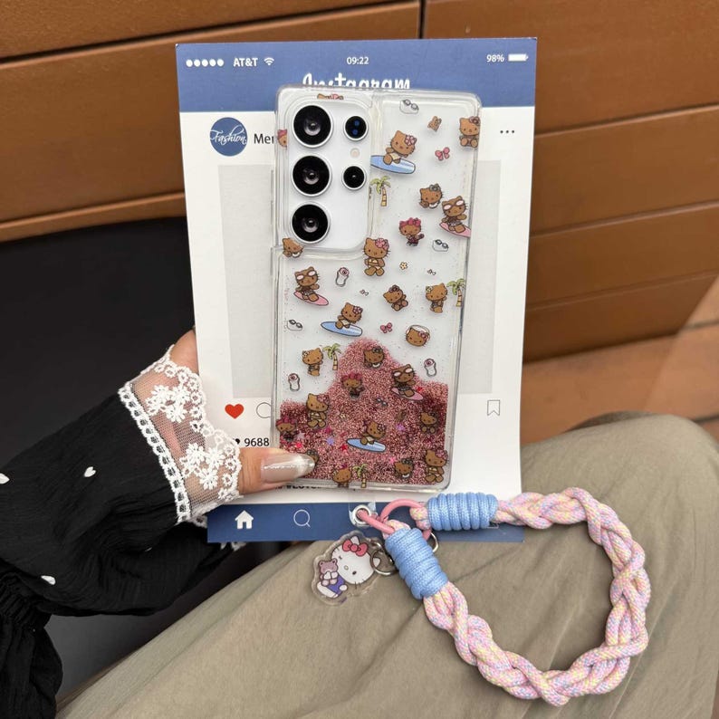 May include: Clear phone case with a Hello Kitty design, featuring surfing characters and pink glitter. Includes a braided wrist strap in pastel colors and a Hello Kitty charm. The phone case is designed for a Samsung Galaxy.