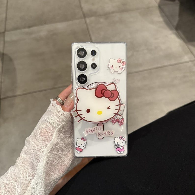 以下が含まれることがあります： ハローキティのデザインが施された透明な携帯電話ケース。ケースには赤いリボンが付いた大きなハローキティの顔があり、その下に「Hello Kitty」の文字が印刷されています。小さなハローキティのグラフィックとリボンも表示されています。