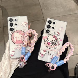 以下が含まれることがあります： ハローキティのデザインが施された透明な携帯電話ケース2つ。各ケースには、ピンクと白の編み込みリストストラップと青いアクセントが付いています。ケースには、さまざまなポーズのハローキティが描かれており、「Hello Kitty」と「Happy day」の文字が書かれています。