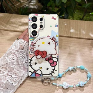May include: A cream-colored phone case featuring a Hello Kitty design with a pop-up grip. The case has a wrist strap with blue beads and charms. The case has a cartoon design with bows, stars, and other decorative elements.