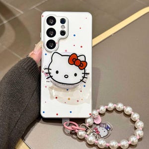 K&ouml;nnte beinhalten: Wei&szlig;es Handycase mit Hello Kitty-Design, roter Schleife und bunten Punkten. Inklusive einer Perlen-Handschlaufe mit Hello Kitty-Anh&auml;nger. Das Handycase hat eine Kameraanordnung.