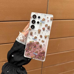 May include: Clear phone case with a beach-themed design. The case features a pattern of brown cartoon characters surfing on blue surfboards, with pink glitter and small clear bubbles. The case has a camera cutout and buttons.