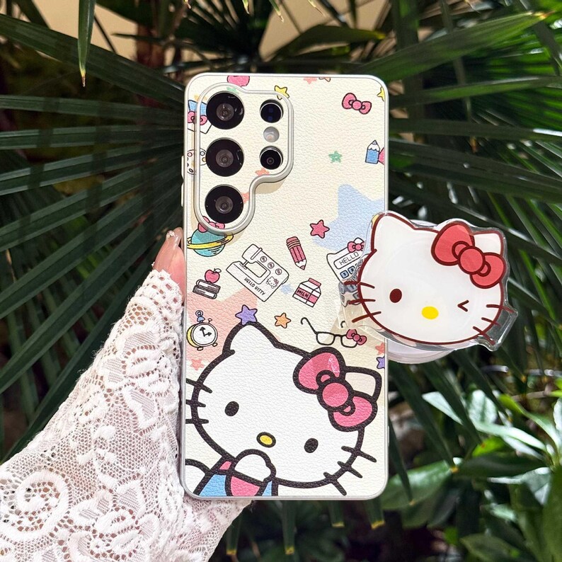May include: White phone case with a Hello Kitty design. The case features a repeating pattern of Hello Kitty, bows, stars, and school supplies. A clear phone grip with a Hello Kitty face is attached to the case.