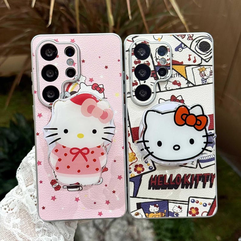 Peut inclure: Deux coques de t&eacute;l&eacute;phone avec des motifs Hello Kitty. L'une est rose avec des &eacute;toiles et un pop-up Hello Kitty. L'autre est blanche avec un motif Hello Kitty et l'inscription "Hello Kitty". Les deux coques ont plusieurs objectifs d'appareil photo.