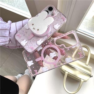 Könnte beinhalten: Zwei transparente Handyhüllen mit rosa Akzenten und einem Hasen-Thema. Eine Hülle hat einen großen weißen Hasen-Pop-up-Griff, die andere einen rosa Kreis mit einem Hasen. Beide Hüllen haben Glitzer und kleine Hasen- und Schleifendesigns.