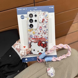 May include: Clear phone case with a Hello Kitty design. The case features a glittery red base with the Hello Kitty character and a variety of other cartoon characters. A pink, blue, and white braided wrist strap is attached to the case.