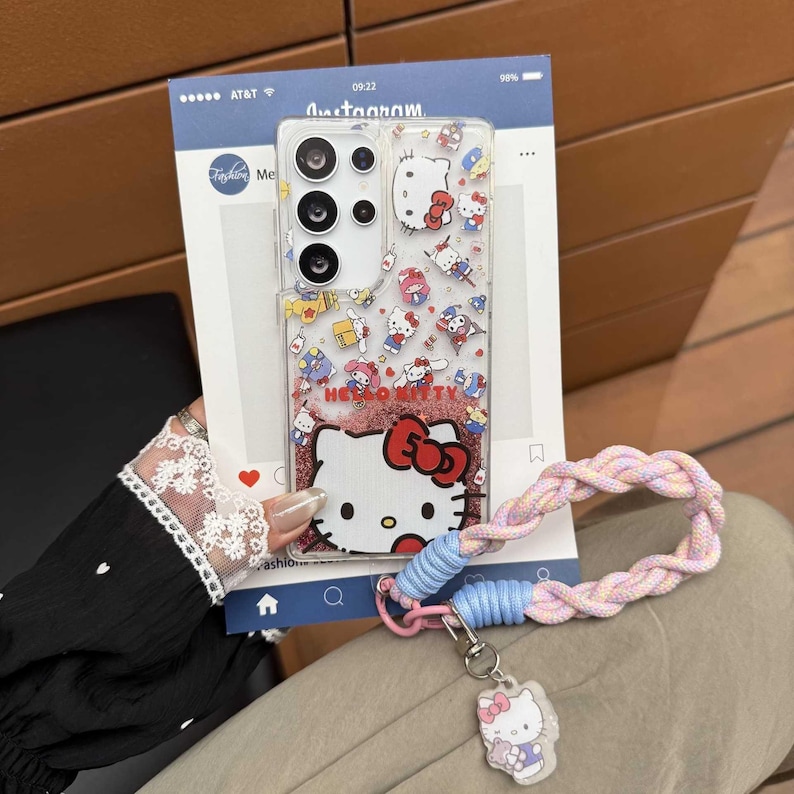 May include: Clear phone case with a Hello Kitty design. The case features a glittery red base with the Hello Kitty character and a variety of other cartoon characters. A pink, blue, and white braided wrist strap is attached to the case.