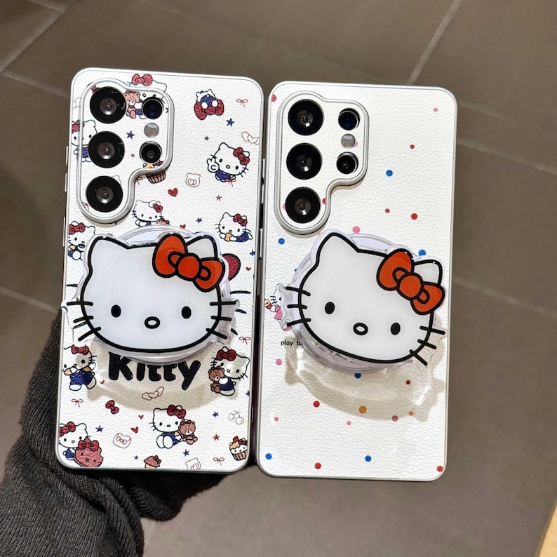 K&ouml;nnte beinhalten: Zwei wei&szlig;e Handyh&uuml;llen mit einem Hello Kitty-Design. Die H&uuml;llen haben einen Pop-up-Griff mit einem Hello Kitty-Gesicht und einer roten Schleife. Die H&uuml;llen sind mit kleinen Hello Kitty-Illustrationen, Herzen und Sternen verziert.