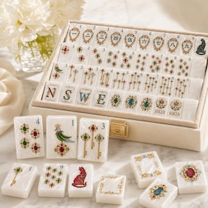 May include: A set of white mahjong tiles with intricate gold, red, green, and blue decorative illustrations. The tiles feature Chinese numerals, cardinal directions, keys, cats, and joker labels, all housed in a beige storage case with a gold latch.