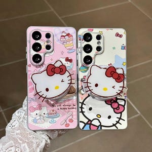 May include: Two phone cases featuring the Hello Kitty character. One case is pink with a dessert and bow design, the other is white with a bow and star design. Each case has a clear ring holder and a Hello Kitty face.
