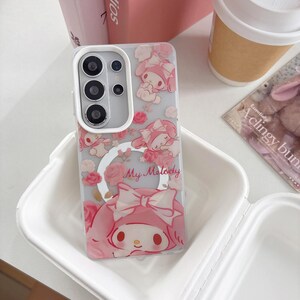 May include: A clear phone case featuring a pink My Melody design with roses and the text "My Melody". The case has a silver ring holder and a camera module with three lenses.